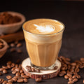 Glass of coffee with latte art surrounded by coffee beans on a dark surface