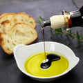 Balsamic vinegar being drizzled into a bowl of olive oil with bread slices on a dark surface.